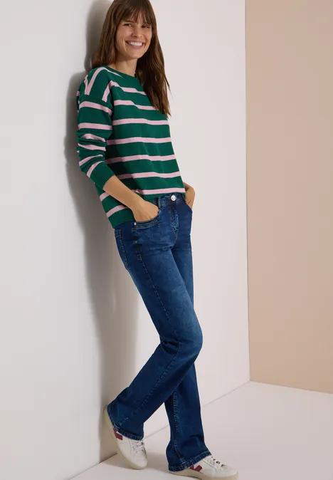 Pull à col bateau et rayures dark fir green melange Femme souriante appuyée contre un mur, pull rayé, jeans et baskets.