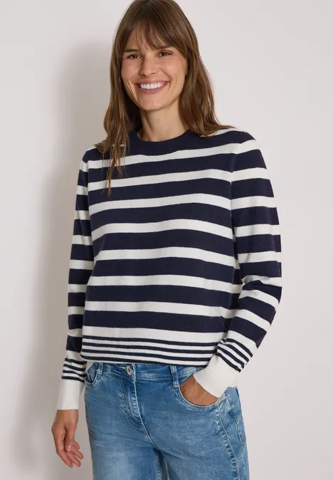 Pull à col rond et à rayures urban dark blue melange Une femme souriante porte un pull à rayures bleu marine et blanc avec un jean devant un mur blanc.
