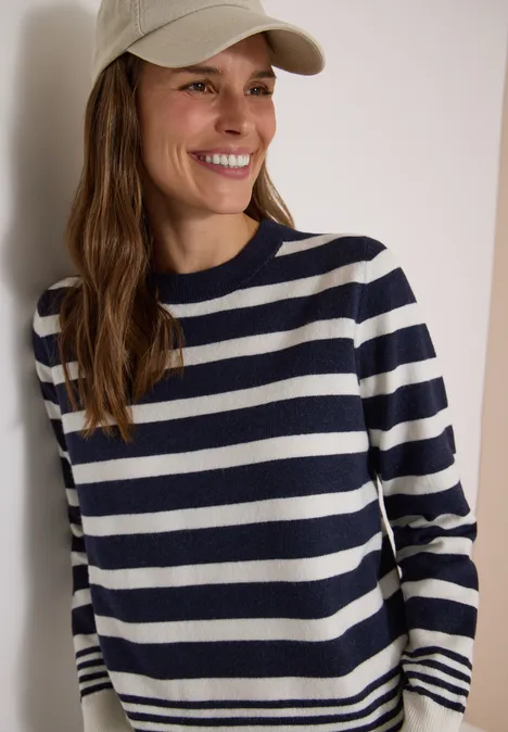 Pull à col rond et à rayures urban dark blue melange Femme souriante portant un pull rayé bleu marine et blanc et une casquette beige devant un fond clair.