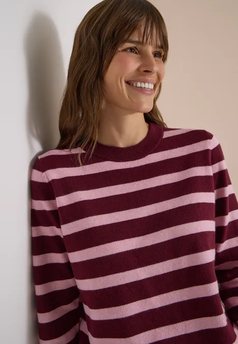 Pull à col rond et rayé red wine melange Femme souriante appuyée sur un mur, vêtue d'un pull rayé bordeaux et rose.