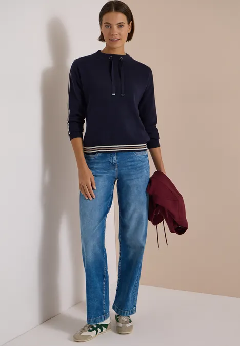 Frau in dunkelblauem Hoodie, Jeans und Sneakern hält eine burgunderrote Jacke.