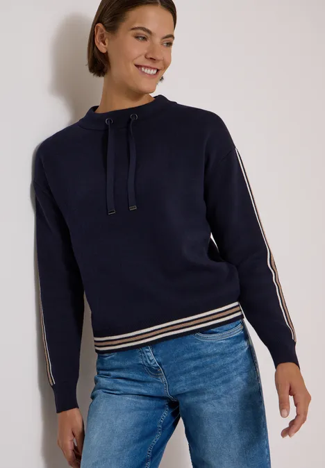 Frau in blauem Kapuzenpullover mit Streifen an Ärmeln und Bund, kombiniert mit Jeans, steht lächelnd.