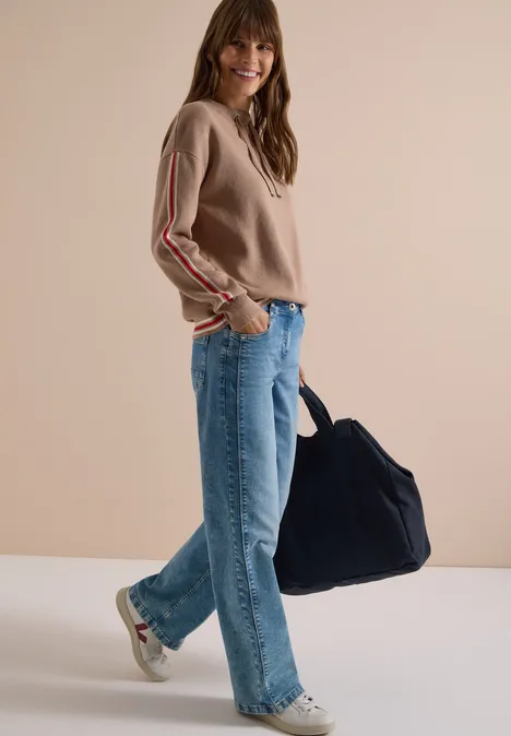 Frau in beigem Pullover, blauen Jeans, Sneakern, trägt große blaue Tasche, vor schlichter Wand lächelnd.