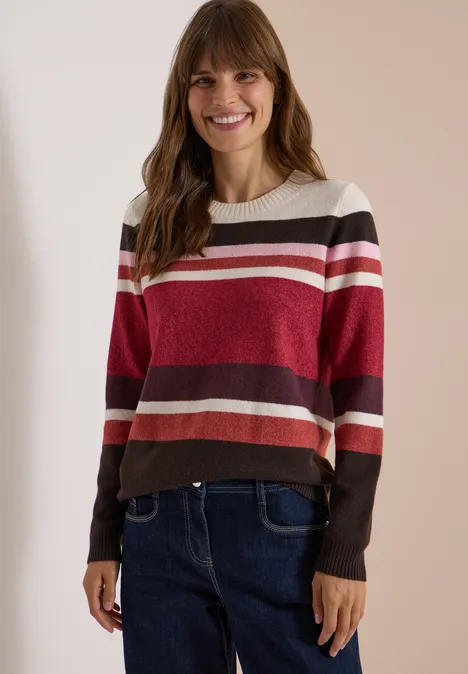 Pull à col rond et rayures rhubarb melange Une femme souriante porte un pull rayé rouge, rose et marron avec un jean, devant un fond clair.