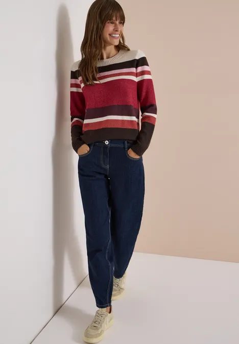 Pull à col rond et rayures rhubarb melange Femme souriante appuyée contre un mur clair, porte un pull rayé, un jean ample et des baskets blanches.