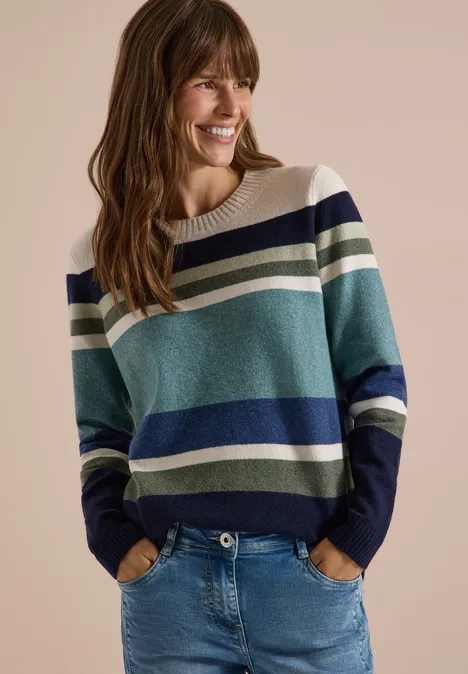 Pull à col rond et à rayures mineral blue melange Femme souriante en pull rayé bleu, vert et blanc avec jean, mains en poches, devant un fond beige.