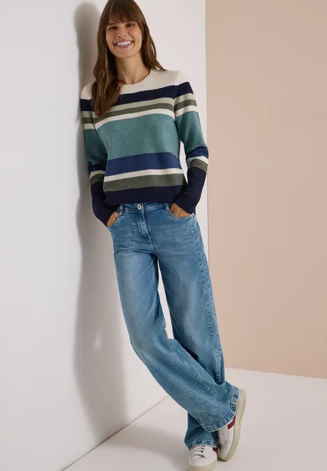 Pull à col rond et à rayures mineral blue melange Une femme souriante appuyée contre un mur, portant un pull rayé, un jean bleu et des baskets.