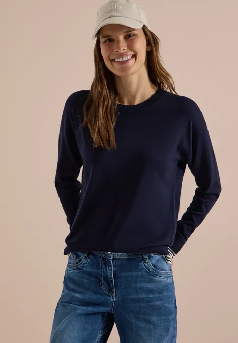 Pull à col rond avec détail superposé urban dark blue Femme souriante en casquette blanche, pull bleu marine et jean, posant de manière décontractée.