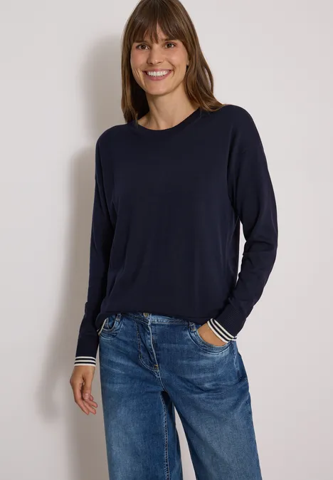 Pull à col rond avec détail superposé urban dark blue Femme en pull bleu foncé à poignets rayés et jeans, souriante devant un fond clair.