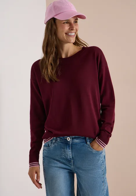 Eine junge Frau mit pinker Cap, bordeauxrotem Langarmshirt und Jeans lächelt entspannt.