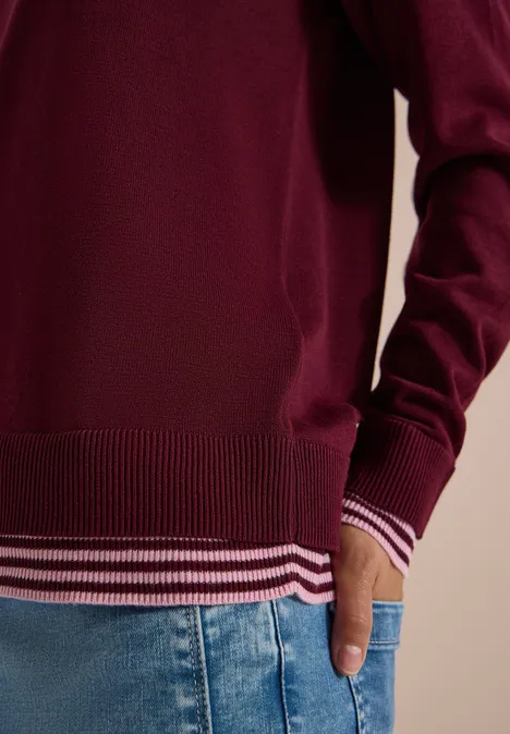 Roter Pullover mit gestreiftem Saum und Jeans, rechte Hand in der Hosentasche.