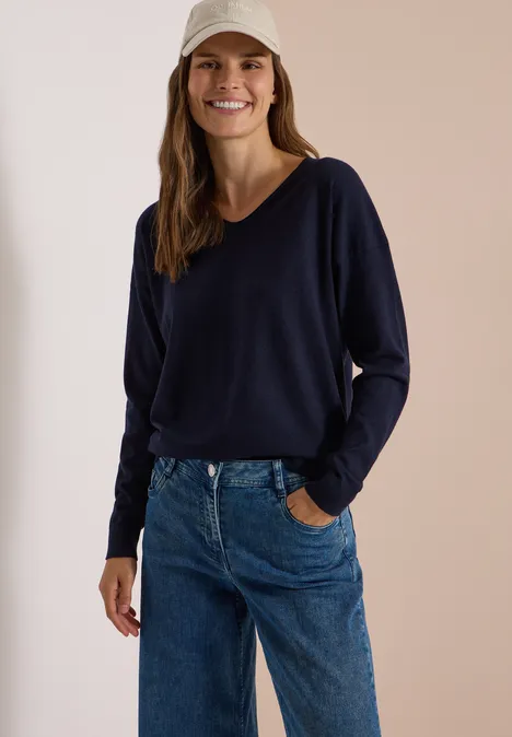 Pull à manches longues et col en V arrondi urban dark blue Femme souriante en pull bleu marine, jean et casquette beige.