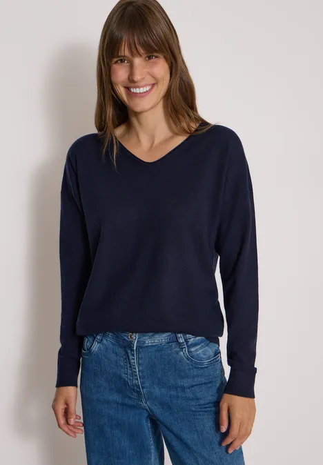 Pull à manches longues et col en V arrondi urban dark blue Une femme souriante en pull bleu foncé et jean devant un fond clair.