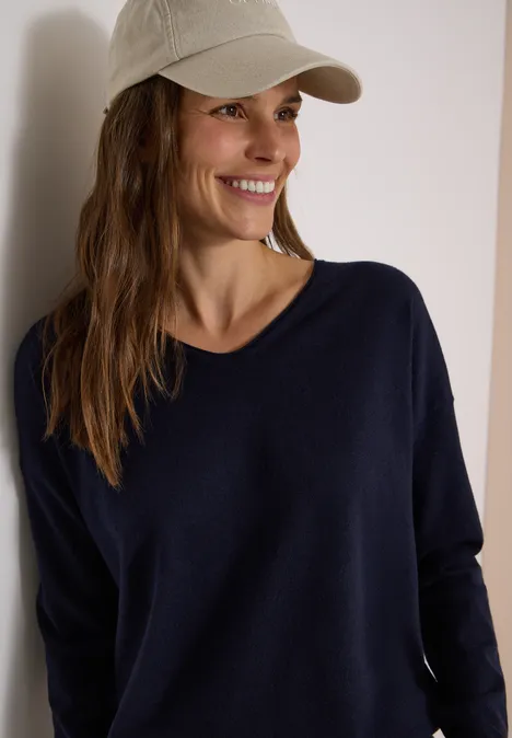 Pull à manches longues et col en V arrondi urban dark blue Femme souriante appuyée sur un mur, casquette beige et pull bleu foncé.