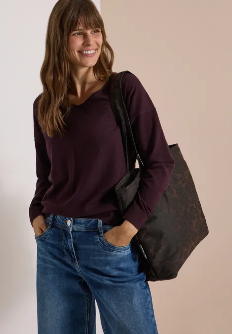 Eine lächelnde Frau trägt einen weinroten Pullover, blaue Jeans und eine große Schultertasche.