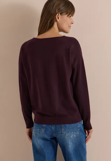 Frau von hinten in lila Pullover und blauen Jeans vor neutralem Hintergrund.