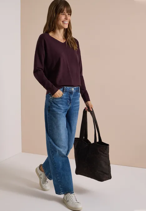 Frau mit braunem Haar in dunklem Pullover, weiten Jeans und weißen Sneakern hält schwarze Tasche.
