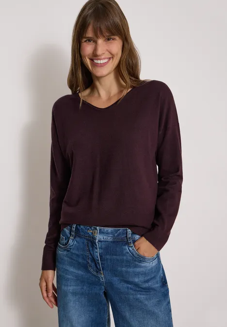 Fröhliche Frau trägt bordeauxfarbenen Pullover und Jeans, posiert lächelnd vor neutralem Hintergrund.