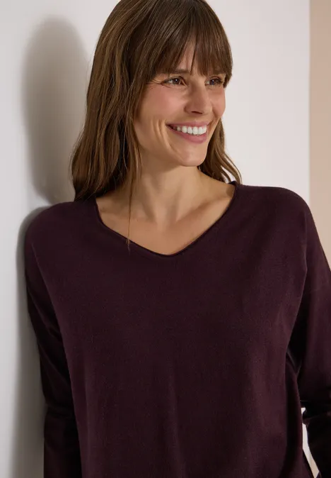 Eine lächelnde Frau in einem pflaumenfarbenen V-Ausschnitt-Pullover, angelehnt an eine weiße Wand.