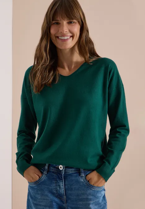 Eine lächelnde Frau in grünem Pullover und Jeans vor neutralem Hintergrund.
