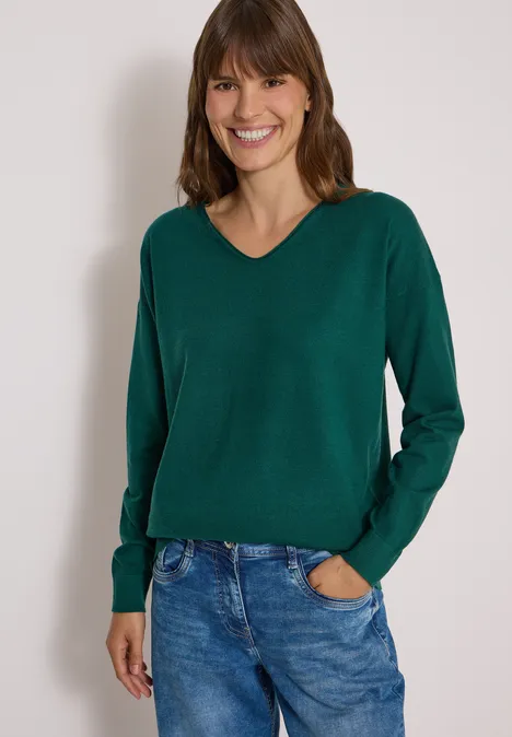 Fröhliche Frau trägt grünen V-Ausschnitt-Pullover und Jeans vor hellem Hintergrund.