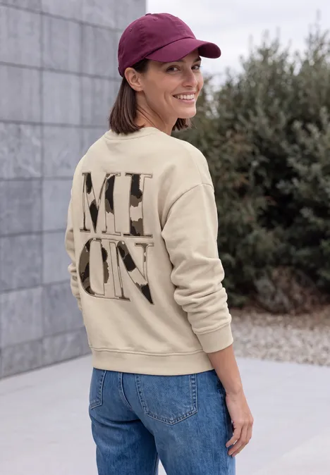 Sweatshirt mit Rundhals und Backprint pearl beige Eine Frau in Jeans und beigem Pullover mit großem Schriftzug auf dem Rücken steht draußen und lächelt zur Kamera.