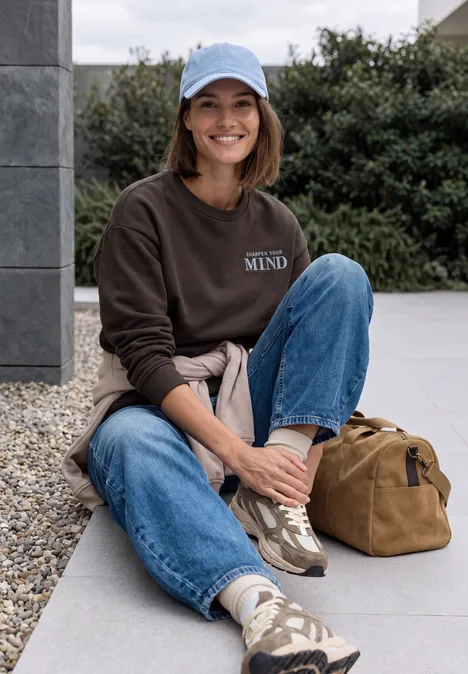 Sweatshirt mit Rundhals und Backprint truffle brown Eine Frau mit Cap, Sweatshirt und Jeans sitzt lachend draußen, Sneakers an den Füßen, Tasche neben sich.