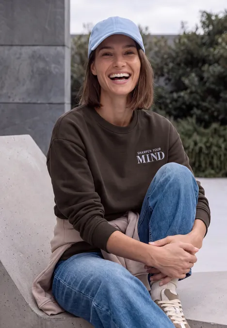 Sweatshirt mit Rundhals und Backprint truffle brown Eine lächelnde Frau im Baseballcap, braunem Sweatshirt und Jeans sitzt im Freien auf einer Bank.