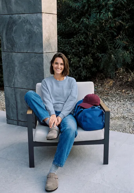 Junge Frau in Jeans und Pullover sitzt auf modernem Stuhl im Freien, lächelt entspannt.