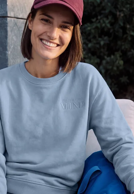 Junge Frau mit roter Baseballkappe und hellem Sweatshirt sitzt lächelnd vor Grünpflanzen.