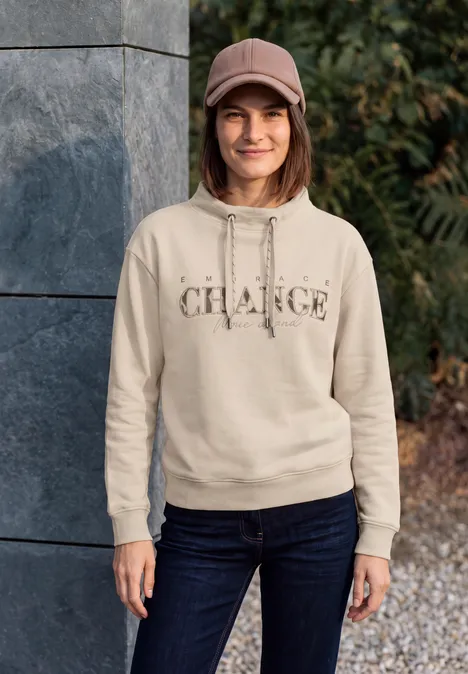 Sweat-shirt avec inscription et col cheminée pearl beige Junge Frau vor Steinmauer trägt hellen Hoodie mit Schriftzug, Jeans und Cap, steht lächelnd im Freien.