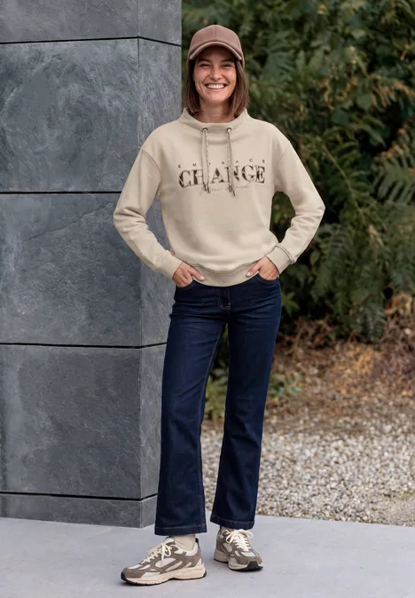 Sweat-shirt avec inscription et col cheminée pearl beige Lächelnde Frau in beigem Hoodie, Jeans und Sneakern steht vor einer Steinwand und grünen Pflanzen.