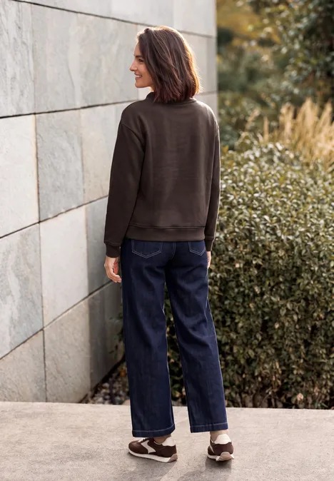 Sweat-shirt avec inscription et col cheminée truffle brown Frau von hinten in lässigem Outfit (Pullover, Jeans, Sneakers) steht vor einer Steinwand und Büschen.