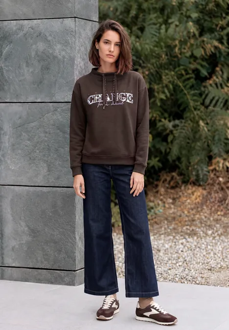 Sweat-shirt avec inscription et col cheminée truffle brown Frau im braunen Kapuzenpullover und weiten Jeans steht vor einer Mauer und grünem Strauchwerk.