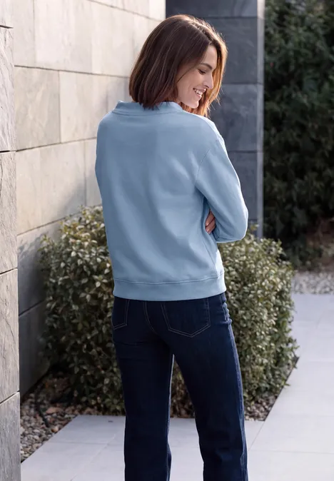 Sweat-shirt avec inscription et col cheminée cosmopolitan blue Frau in blauem Pullover und Jeans lehnt seitlich an Hauswand, Heckengebüsch im Hintergrund.