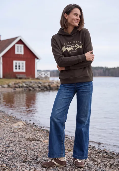 Vrouw in hoodie en jeans glimlachend aan de oever van een meer, met een rood huisje op de achtergrond.