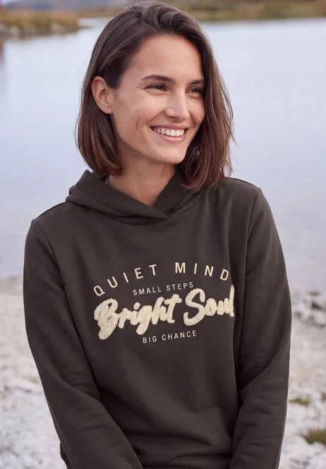 Vrouw glimlachend in hoodie aan de oever van een meer, tekst op de voorkant.
