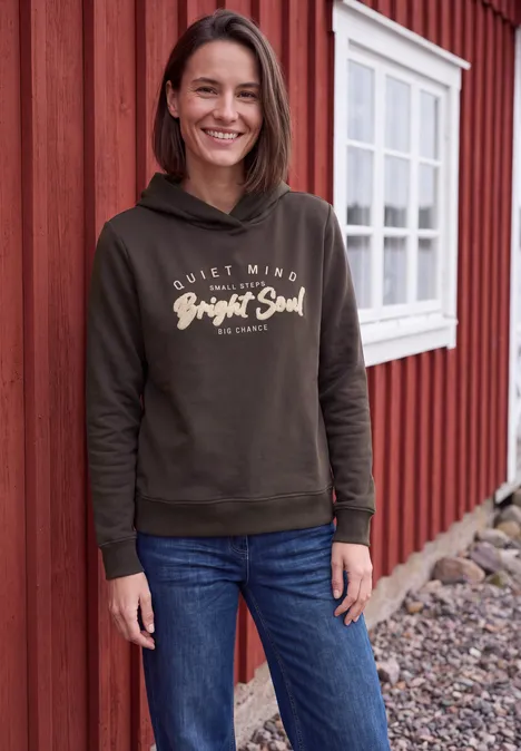 Een lachende vrouw voor een rood houten huis, draagt jeans en een hoodie met 'Bright Soul'.