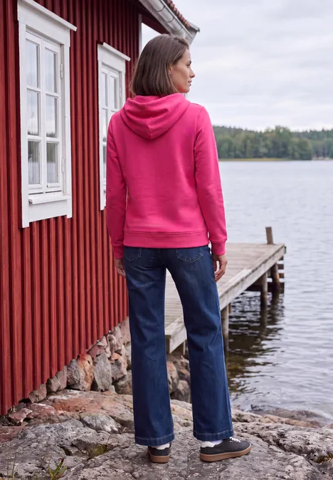 Sweatshirt mit Details in Rippstruktur pink flash Frau in pinkem Kapuzenpullover und Jeans steht am Ufer neben roter Holzhütte und blickt auf den See.