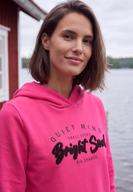 Sweatshirt mit Details in Rippstruktur pink flash Junge Frau in pinkem Hoodie steht lächelnd am Seeufer vor rotem Holzhaus.