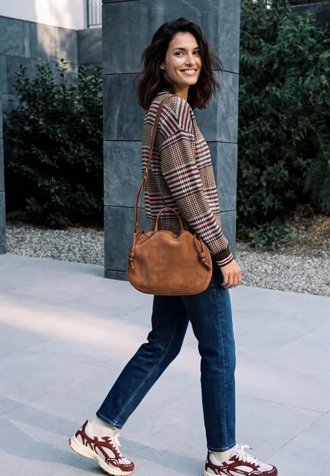 Eine lächelnde Frau in Jeans, kariertem Pullover und Sneakers trägt eine braune Tasche vor modernem Gebäude.