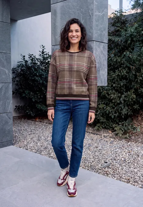 Eine lächelnde Frau trägt Pullover und Jeans und steht vor einer Mauer mit Pflanzen.