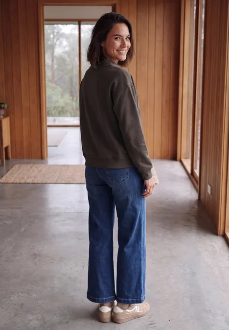 Een vrouw in een groene jas en blauwe jeans staat glimlachend om zich heen te kijken in een lichte houten hal.