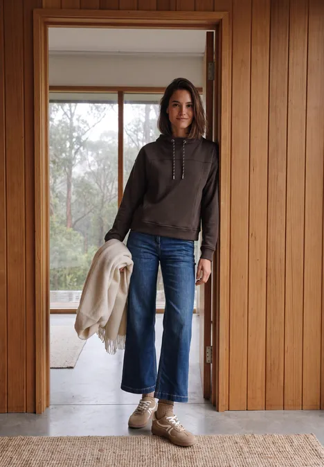 Vrouw in spijkerbroek en hoodie, jas in hand, staand in een houten kozijn voor een raam.
