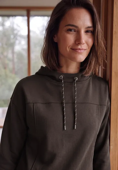 Een vrouw met bruin haar draagt een donker hoodie en glimlacht voor een raam.