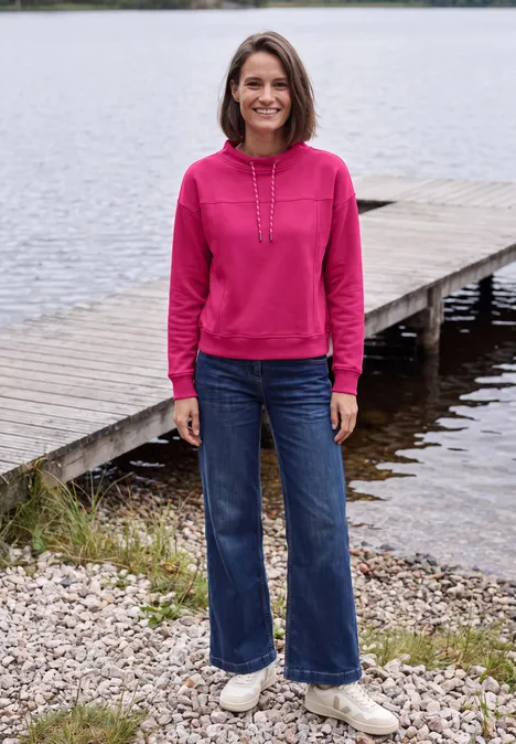 Een vrouw in een roze hoodie en spijkerbroek staat lachend op een houten steiger bij het meer.