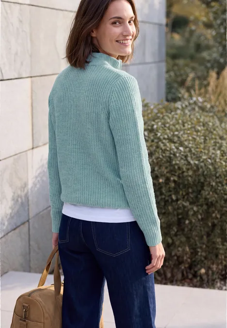 Frau in mintgrünem Pullover und Jeans hält braune Tasche, steht lächelnd vor Mauer und Büschen.