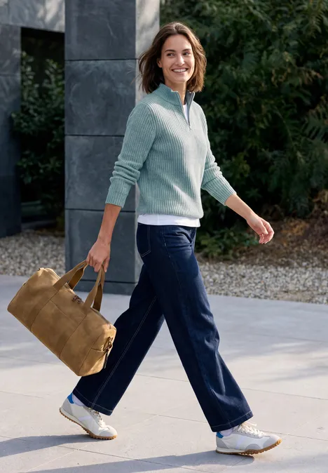 Frau spaziert draußen, lächelnd, trägt blaue Hose, hellblauen Pullover und eine braune Wildledertasche.