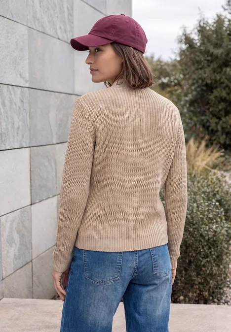 Pullover in Unifarbe mit Zipper creamy mocha melange Frau steht seitlich vor einer Steinmauer, trägt rote Kappe, beigen Pullover und blaue Jeans.