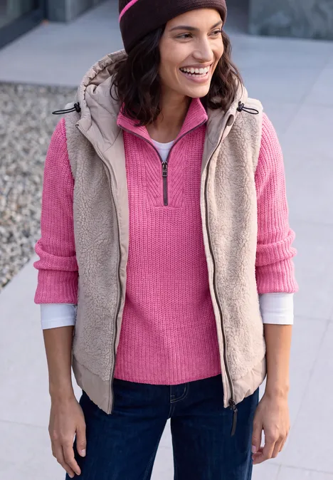 Trui met rits pink flash melange Frau in Jeans mit rosa Pullover, beige Weste und Mütze lächelt im Freien.